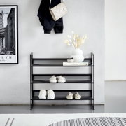 Schwarzes Metall-Schuhregal mit drei Ablagen im Flur, frontal in leichter Untersicht fotografiert, mit mehreren Paar Schuhen, Deko-Vase und Buch auf der oberen Ebene vor heller Wand